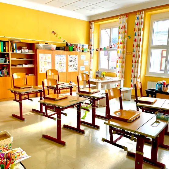 Ein Klassenzimmer mit gelben Wänden und weißen Rahmen ist mit mehreren Holztischen und Stühlen ausgestattet. Eine bunte Banner mit Buchstaben schmückt die Wand. Es gibt Regale mit Büchern und Aufbewahrungsboxen. Zwei Fenster mit Vorhängen sorgen für natürliches Licht.