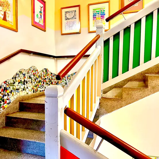 Eine Treppe mit mehrfarbigen Fliesen und Holzhandläufen. Die Stufen haben gelbe, grüne und rote Streifen. Über den Stufen hängen drei Bilderrahmen an der Wand.