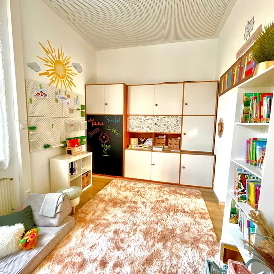 Ein Zimmer mit einem Sonnendesign an der Wand und einem rosa Teppich auf dem Boden. Der Raum hat Schränke und Regale voller Bücher. Es gibt ein Sofa mit einer Decke und Kissen.