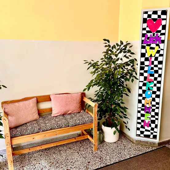 Eine gemütliche Holzbank mit zwei rosa Kissen und einem Topfpflanzen steht neben einem bunten Schild, das 'Ich will komme' besagt, an einer gelben und weißen Wand.