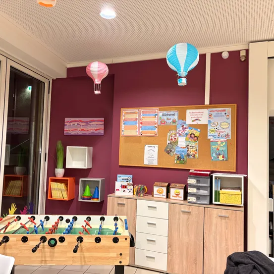 Ein Raum mit einer roten Wand, einer Pinnwand und einem Tischfußball. Eine Glastür führt in den Garten, und verschiedene Dekorationen schmücken den Raum.