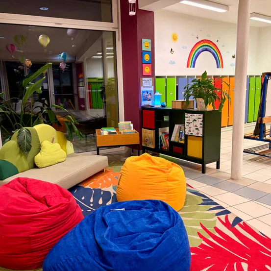 Ein farbenfroher Kinderzimmer mit Beanbags, leuchtenden Möbeln und bunten Spinden. Ein Regenbogen und eine Sonne sind an der Wand gemalt.