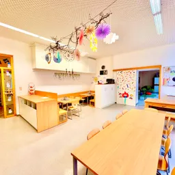 Ein leerer Klassenraum mit langen Holztischen und Stühlen. Ein weißer Schrank und eine Tür sind sichtbar. Dekorationen hängen von der Decke.