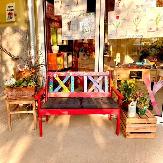 Eine rote Bank mit farbenfrohen Designs steht auf dem Bürgersteig vor einem Geschäft mit Blumenladen-Display und Lehrtafeln.