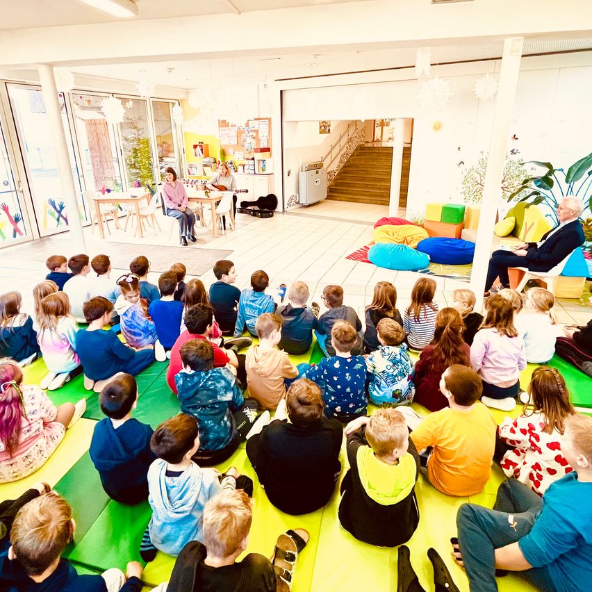 Ein Klassenzimmer mit Kindern auf dem Boden sitzend, zwei Erwachsene stehen in der Mitte. Die Erwachsenen sitzen auf Stühlen, und eine Treppe ist hinter ihnen. Ein Tisch mit Stühlen ist auf der linken Seite. Ein Teppich mit Kissen liegt auf dem Boden.