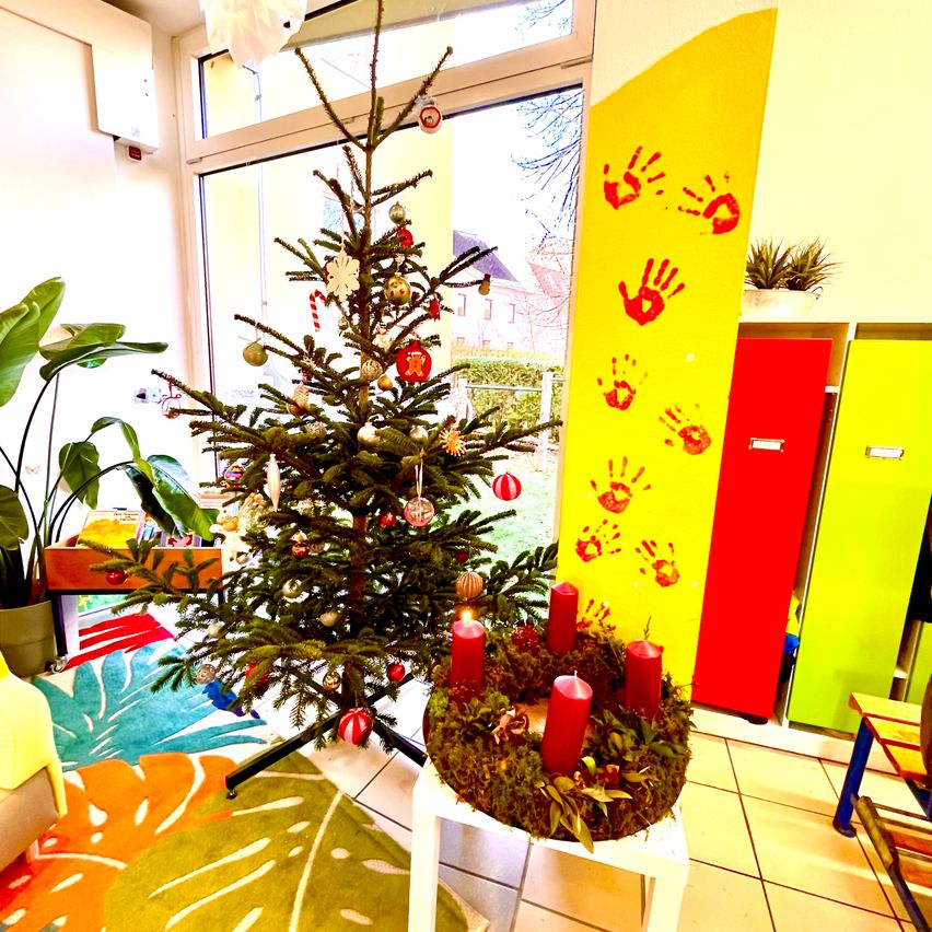 Ein Weihnachtsbaum mit Ornamenten steht in einem Raum mit einer gelben Wand, die mit Handabdrücken verziert ist. In der Nähe steht ein Kranz mit roten Kerzen auf einem weißen Tisch.
