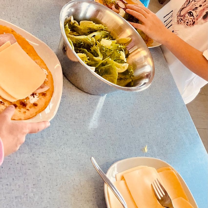 Ein Tisch mit zwei Händen, die ein Sandwich und eine Schüssel Salat halten. Eine Gabel und gefaltete Servietten liegen auf einem anderen Teller. Ein Leopardenmuster-T-Shirt ist teilweise zu sehen.