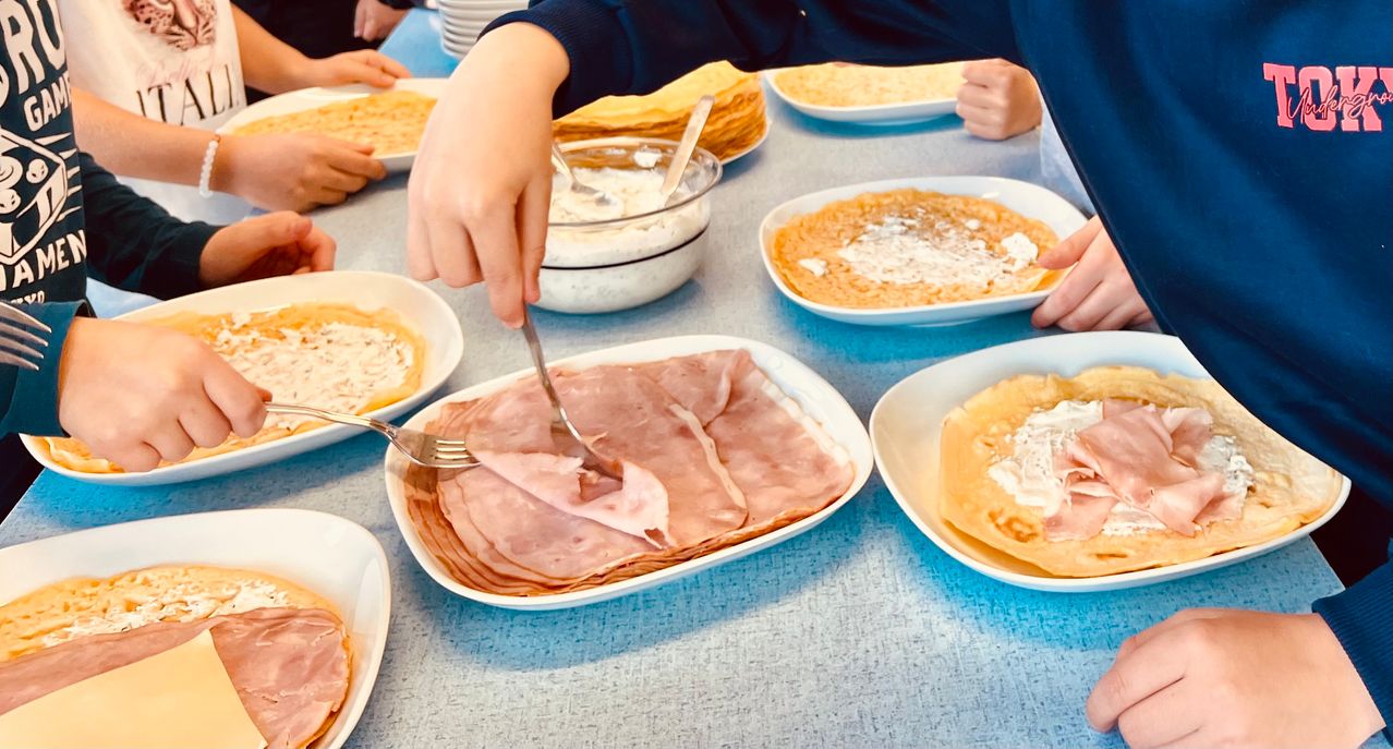 Eine Person schneidet Schinken auf einem Teller, umgeben von anderen Tellern mit Essen. In der Nähe sind Schalen mit Käse und ein Stapel Pfannkuchen zu sehen.