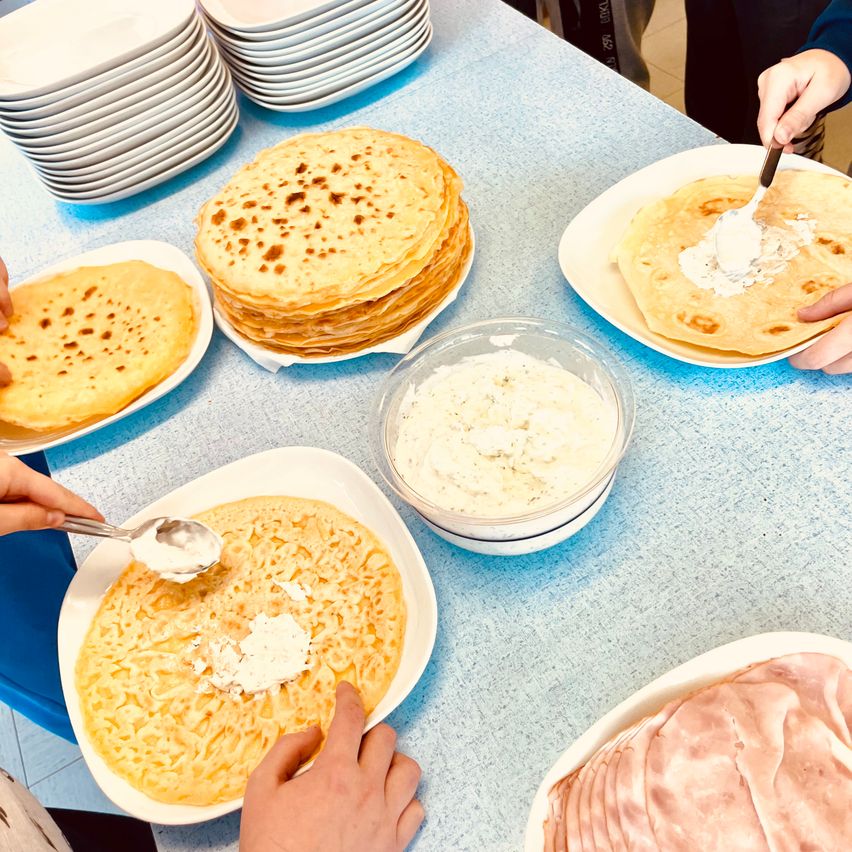 Ein blauer Tisch zeigt verschiedene Teller mit Crêpes, eine Schüssel mit weißer Sauce und Schinkenscheiben. Menschen bereiten die Crêpes mit der Sauce zu.