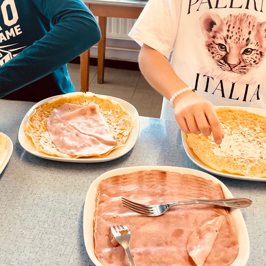 Zwei Personen bereiten ein Gericht zu, wobei eine Person Schinken auf einen Crêpe legt. Das Gericht ist mit Käse überbacken. Ein anderer Teller hat eine Gabel auf einem Fleischgericht.