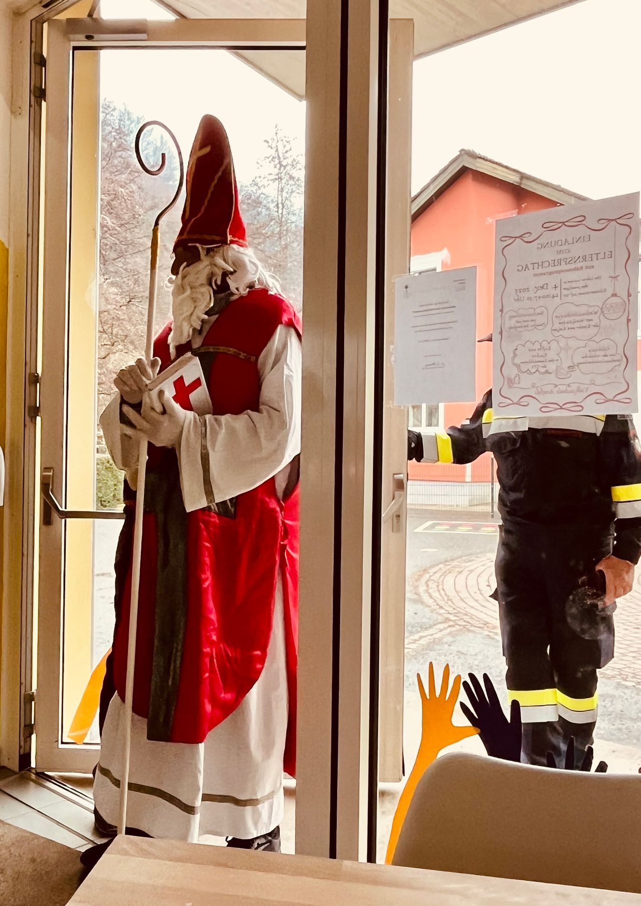 Eine Person in der Kleidung des Heiligen Nikolaus hält ein Buch mit einem roten Kreuz in der Hand und steht neben einer Glastür. Ein Feuerwehrmann in Uniform steht vor der Tür.