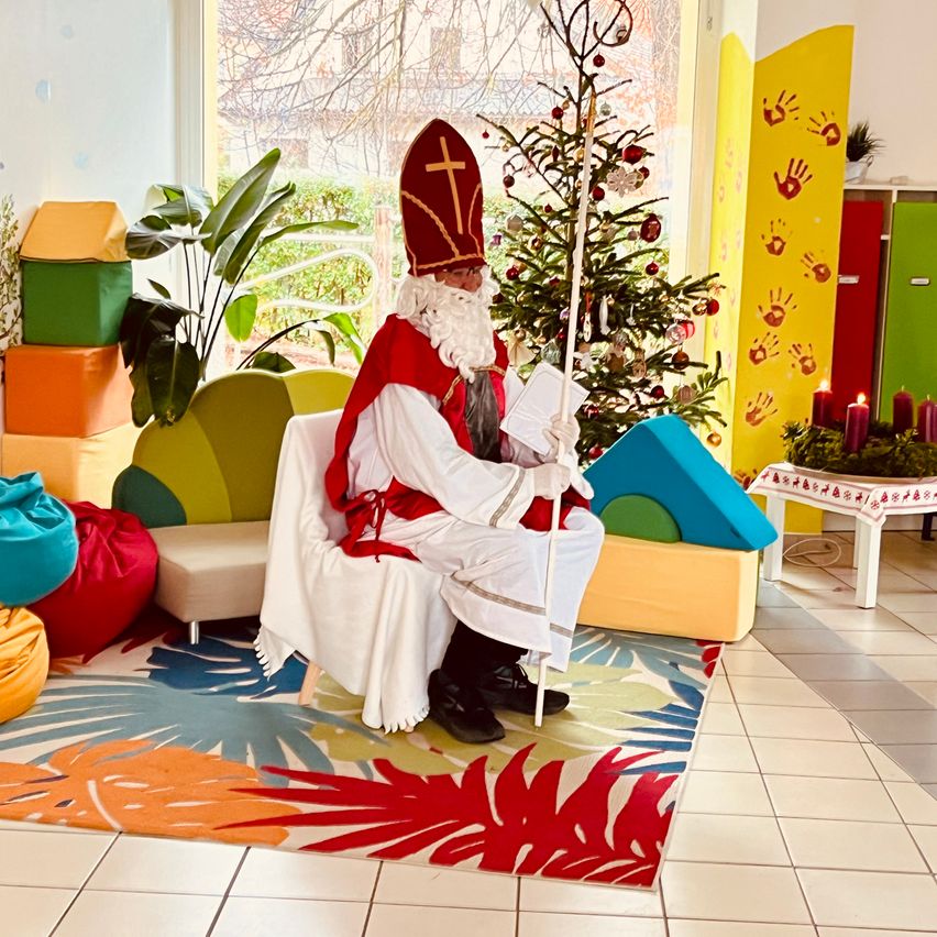 Eine als Weihnachtsmann verkleidete Person sitzt auf einem Stuhl in einem farbenfrohen Raum mit Pflanzen und einem Weihnachtsbaum.