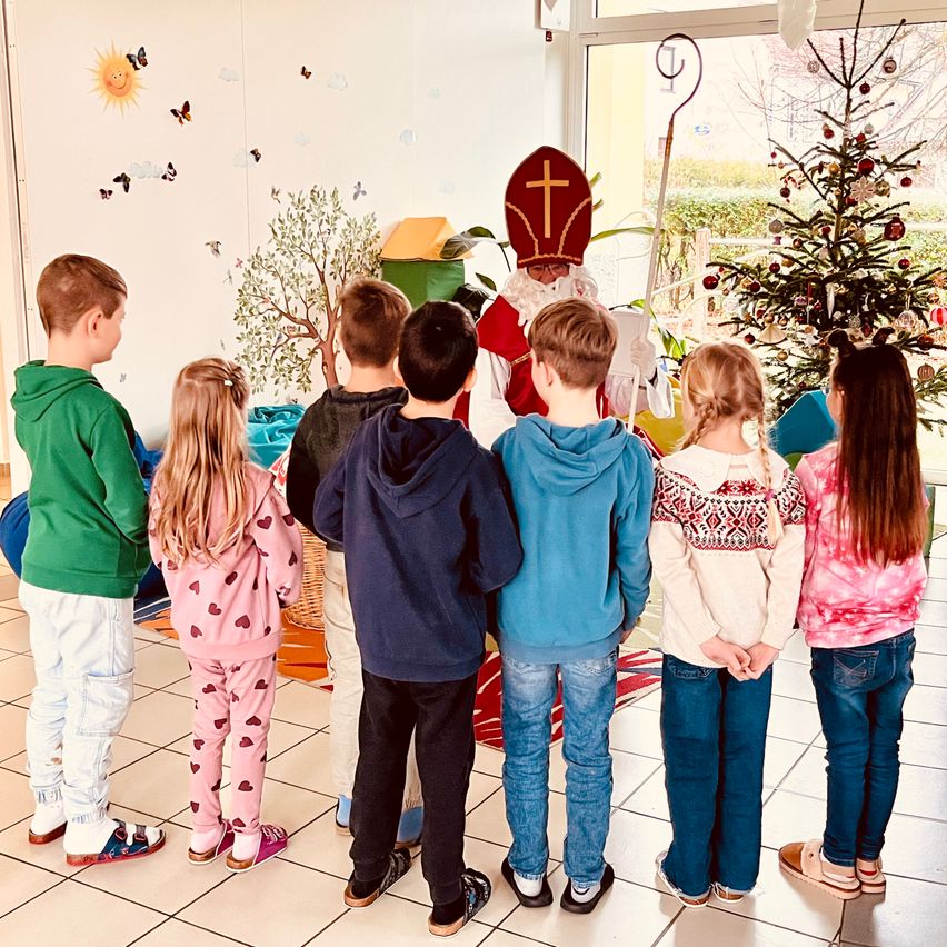 Eine Gruppe von Kindern steht im Kreis und sieht einem Mann zu, der als Weihnachtsmann gekleidet ist. Hinter ihm ist durch eine Glastür ein Weihnachtsbaum zu sehen. An der Wand hinter den Kindern befindet sich ein Wandbild mit Schmetterlingen und einer Sonne.