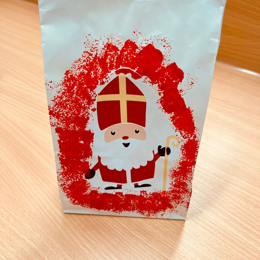 Ein weißer Papierbeutel mit roten Spritzern zeigt einen fröhlichen Weihnachtsmann, der einen Bischofsstab hält. Er trägt einen roten Umhang und ein goldenes Kreuz auf seiner Bischofsmütze.