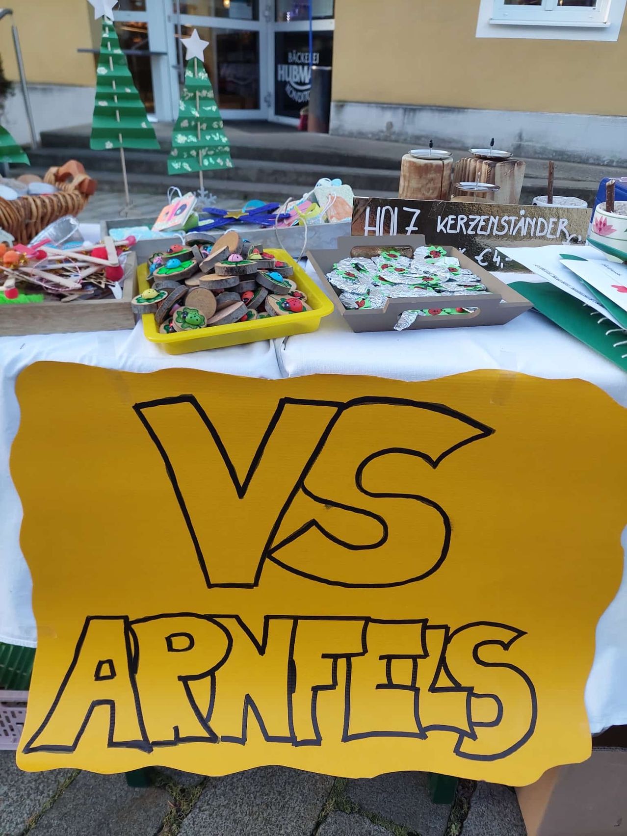 Ein Tisch mit verschiedenen Handwerksarbeiten, Kerzen und selbstgemachten Gegenständen ist ausgestellt. Ein gelbes Schild mit schwarzen Schriftzeichen sagt 'VS ARNTEIS'. Dahinter befinden sich Stufen, ein kleiner Baum und ein Korb.