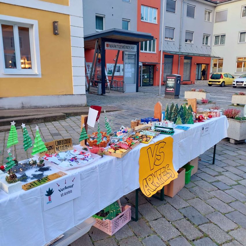 Ein Tisch mit weißem Tuch zeigt handgefertigte Weihnachtsdekorationen, kleine Bäume und Bastelarbeiten. Er steht in einem öffentlichen Bereich mit Gebäuden im Hintergrund. Ein gelbes Schild mit dem Text 'VS ARKETS' ist auf dem Tisch.