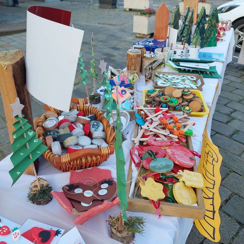 Ein Tisch mit einer weißen Tischdecke zeigt verschiedene handgefertigte Gegenstände, darunter Kekse, Ornamente und einen Korb mit Artikeln. Der Tisch steht draußen auf einem Kopfsteinpflasterweg.