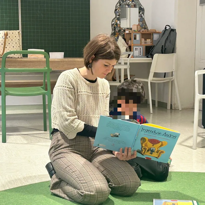 Eine Frau liest einem Kind in einem Zimmer mit grünem Teppich und grünen Stühlen ein Buch vor. Das Buch heißt Irgendwie Anders.
