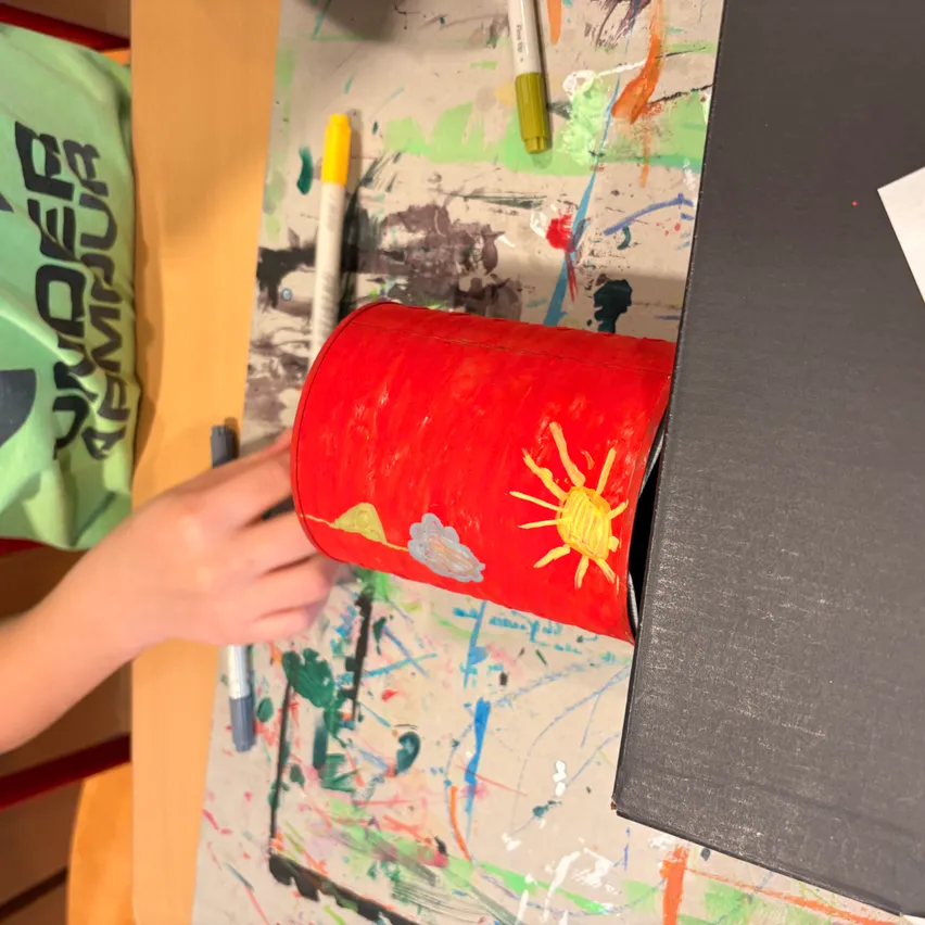 Ein Kind hält eine rot bemalte Röhre, wahrscheinlich ein Bastelprojekt, vor einer Wand mit lebhaften Farbspritzern. Ein Marker und eine Schachtel sind in der Nähe.