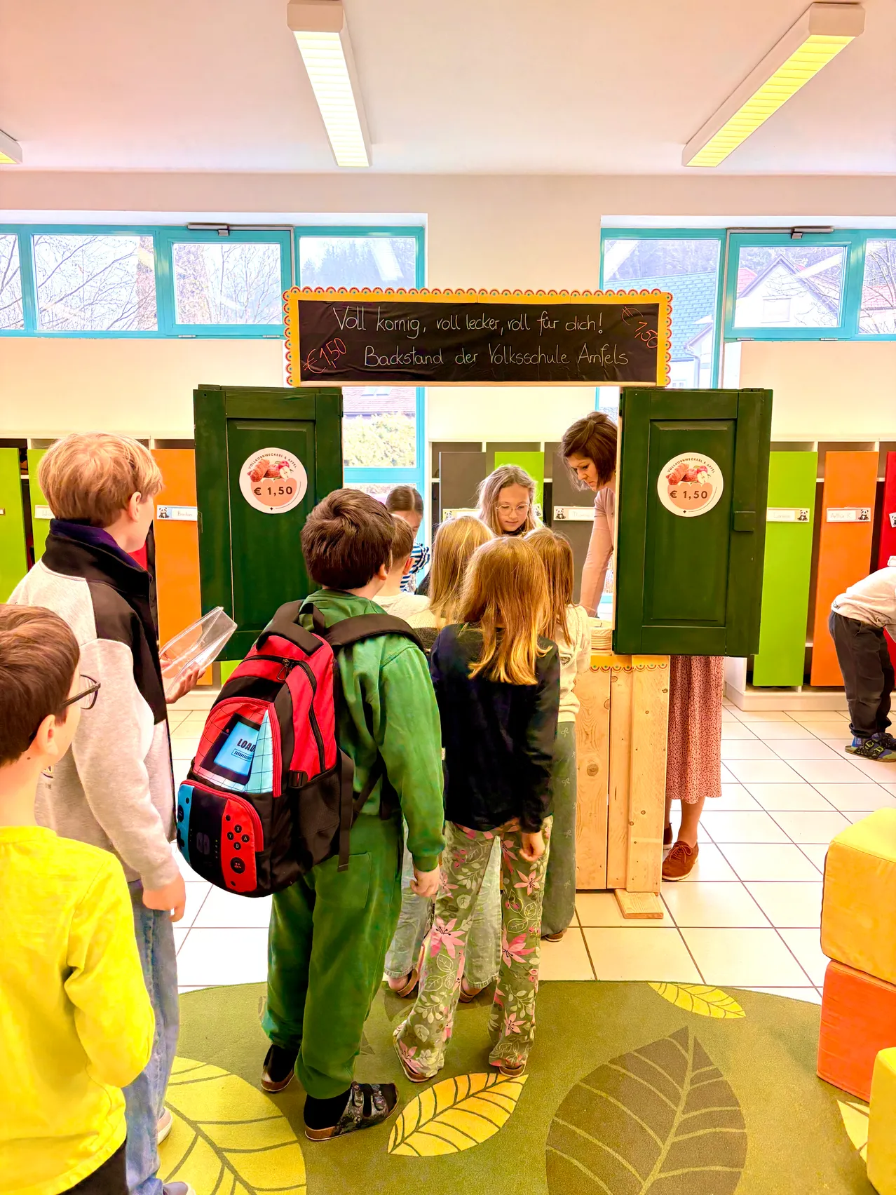 In einem Klassenzimmer steht eine Frau hinter einem Stand mit einer Tafel. Kinder versammeln sich um sie, einige tragen Brillen. Eine grüne Reihe von Schließfächern befindet sich hinter ihnen, und farbige Schließfächer sind auf beiden Seiten.