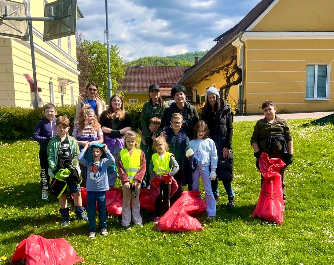 Eine Gruppe von Kindern und Erwachsenen posiert auf einem Grasfeld mit roten Müllsäcken für ein Foto vor gelben Gebäuden.
