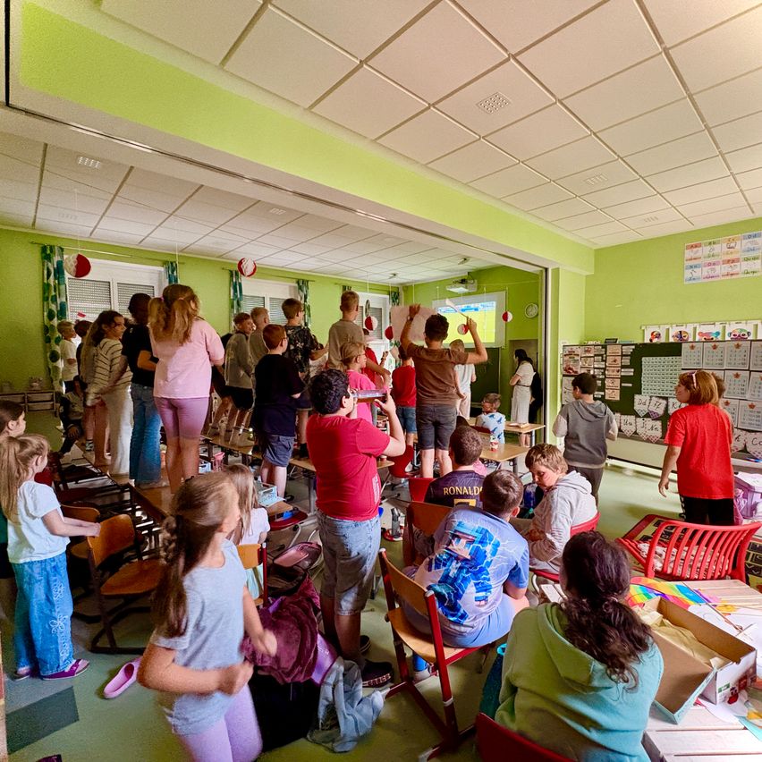 Ein Klassenzimmer mit grünen Wänden, Schüler stehen und sitzen, eine Person benutzt einen Projektor. Eine Uhr und eine Pinnwand sind an der Wand. Rote Stühle sind verstreut.