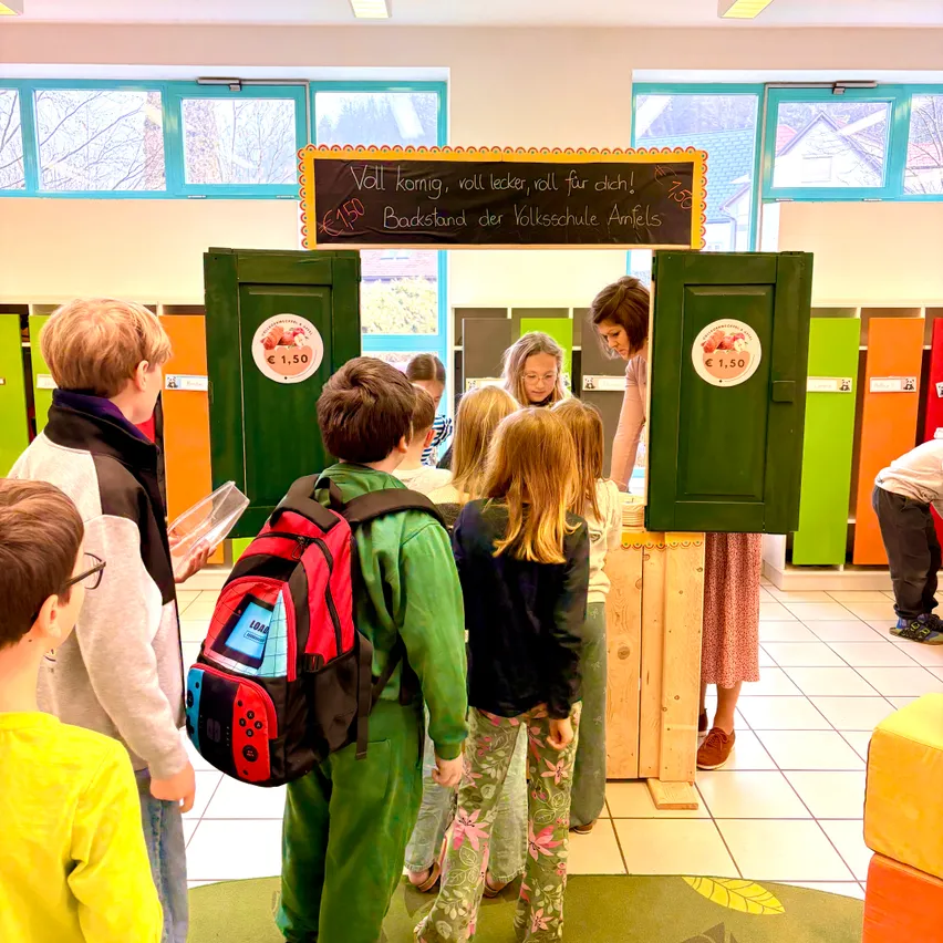 Kinder in einem Klassenzimmer stehen um einen grünen Obststand herum. Über dem Stand hängt ein schwarzes Brett mit der Aufschrift 'Voll komig, voll lecker, roll für dich!'. Die Kinder betrachten die Früchte und einer hält einen Rucksack.
