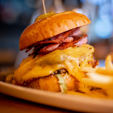 Ein Nahaufnahme eines Hamburgers mit geschmolzenem Käse, Speck und einer Essiggurke sitzt auf einem weißen Teller mit Pommes.
