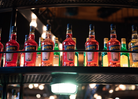 Ein Regal mit mehreren Aperol-Flaschen, die in einer Reihe angeordnet sind, jede mit einem blauen Etikett mit dem Wort Aperol. Die Flaschen werden von einem Licht darunter beleuchtet.