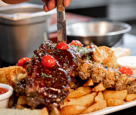 Ein großer Teller mit Rippchen, üppig mit Barbecue-Sauce garniert und mit Kirschtomaten und Kräutern verziert. Dazu gehören Brot, Pommes und Beilagen. Eine Person schneidet die Rippchen mit einem Messer.
