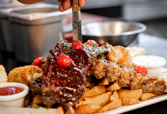 Ein großer Teller mit Rippchen, üppig mit Barbecue-Sauce garniert und mit Kirschtomaten und Kräutern verziert. Dazu gehören Brot, Pommes und Beilagen. Eine Person schneidet die Rippchen mit einem Messer.