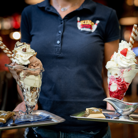 Eine Kellnerin hält zwei Eisbecher auf Tabletts. Der linke ist Schokolade mit Nüssen und der rechte ist Erdbeere mit Eiscreme.