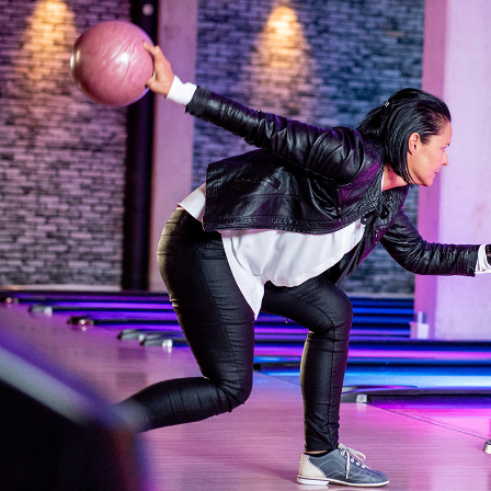 Eine Frau in einer schwarzen Lederjacke bowlt. Sie hält einen rosa Bowlingball, bereit zum Werfen.