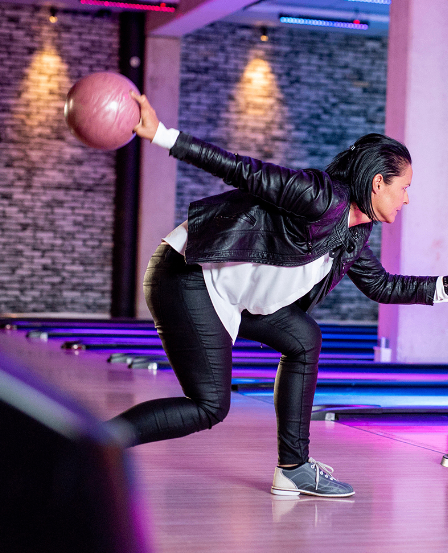 Eine Frau in einer schwarzen Lederjacke bowlt. Sie hält einen rosa Bowlingball, bereit zum Werfen.