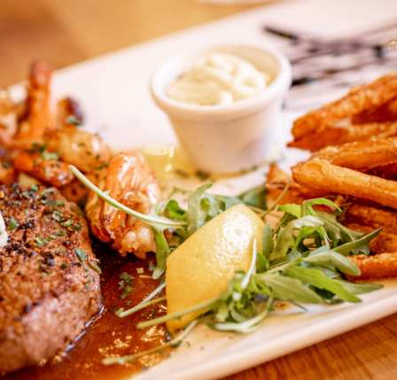 Ein Teller mit Steak, Garnelen und Pommes Frites, garniert mit Zitrone und Kräutern, serviert mit einer kleinen Schüssel cremiger Sauce.