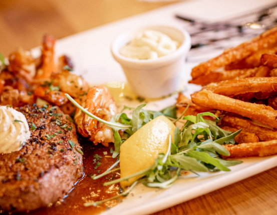 Ein Teller mit Steak, Garnelen und Pommes Frites, garniert mit Zitrone und Kräutern, serviert mit einer kleinen Schüssel cremiger Sauce.