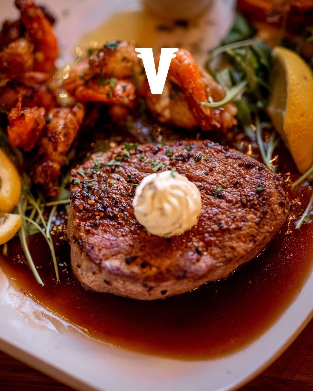 Ein Teller mit Steak und Sauce, garniert mit Kräutern, begleitet von einer Zitronenscheibe und anderen Gerichten mit Garnelen und Karotten.