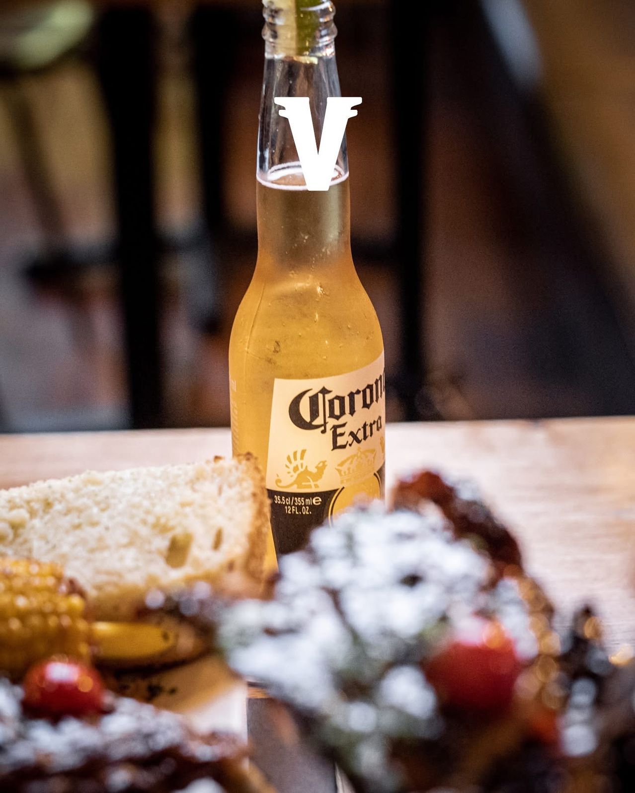 Eine Flasche Corona Extra Bier steht auf einem Holztisch mit einer Scheibe Brot und verschiedenen Lebensmitteln im Hintergrund.