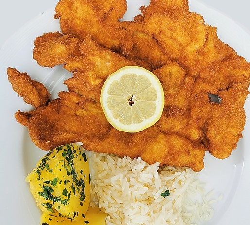 Ein Teller mit gebratenem Fisch, einer Zitronenscheibe und Reis mit Beilage, auf einem weißen Teller serviert.