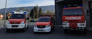 Drei Feuerwehrfahrzeuge, eines mit einem roten Tarp, sind vor einer Feuerwehrstation geparkt, wo eine Person steht.