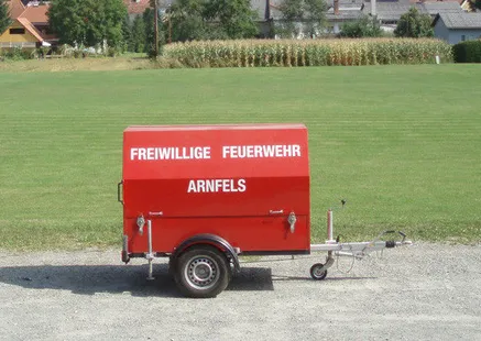 Ein roter Anhänger der Freiwilligen Feuerwehr Arnfels steht auf einem Kiesweg. Im Hintergrund ist ein Rasenfeld und einige Häuser zu sehen.