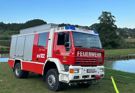 Ein rot-weißer Feuerwehrwagen mit 'Feuerwehr' und 'TLF-A 4000' auf der Seite steht auf dem Gras in der Nähe eines Gewässers.