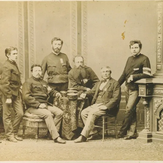 Ein altes Foto zeigt eine Gruppe von Männern in Uniform, einige sitzend und andere stehend. Die Umgebung erscheint formell mit einem dekorativen Kamin im Hintergrund.