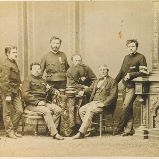 Ein altes Foto zeigt eine Gruppe von Männern in Uniform, einige sitzend und andere stehend. Die Umgebung erscheint formell mit einem dekorativen Kamin im Hintergrund.