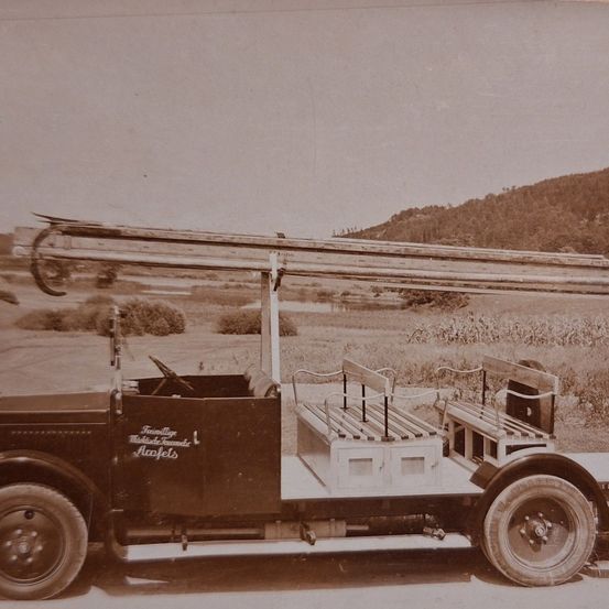 Ein altes Foto zeigt ein Vintage-Fahrzeug mit einer großen Leiter auf dem Dach. Es steht auf einem weitläufigen Feld in einer ländlichen Gegend mit Hügeln im Hintergrund.