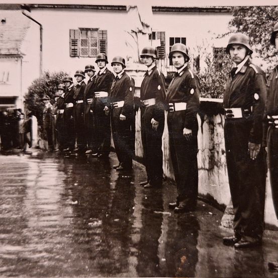 Eine Gruppe von Männern in Militäruniformen steht in einer Reihe vor einem Gebäude auf einer nassen Straße.