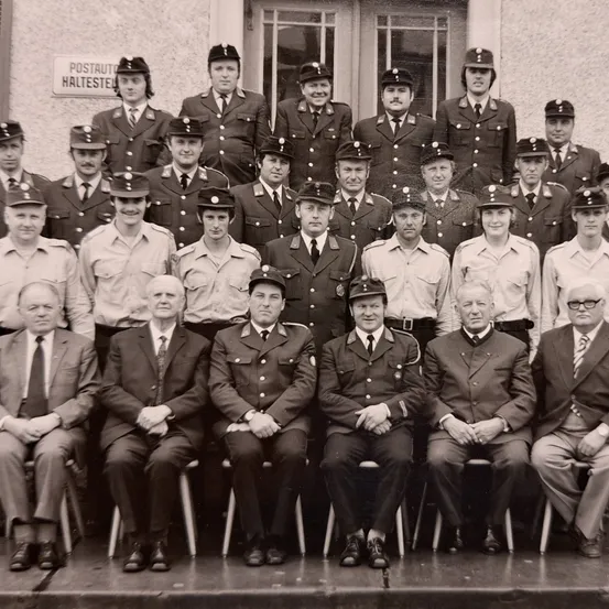 Ein Gruppenfoto von uniformierten Personen, möglicherweise Feuerwehrleute, posiert vor einem Gebäude mit einem Schild, das 'Postauto Haltestelle' liest. Das Bild zeigt eine Mischung aus stehenden und sitzenden Mitgliedern.