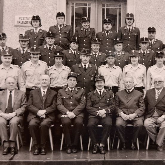Ein Gruppenfoto von uniformierten Personen, möglicherweise Feuerwehrleute, posiert vor einem Gebäude mit einem Schild, das 'Postauto Haltestelle' liest. Das Bild zeigt eine Mischung aus stehenden und sitzenden Mitgliedern.
