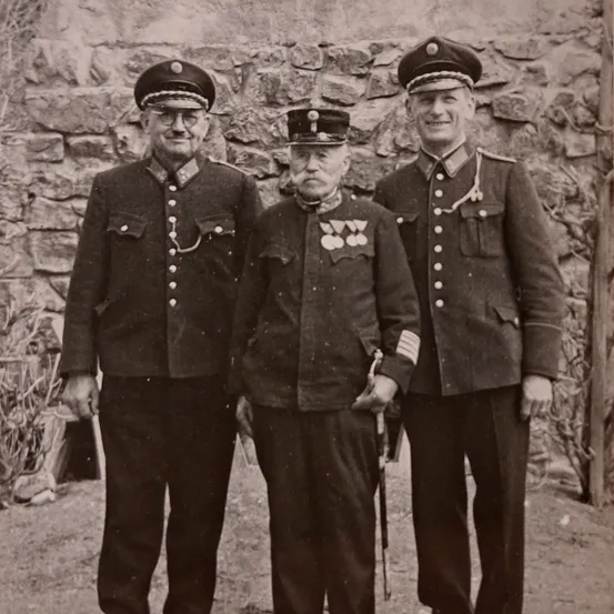 Drei Männer in Uniform stehen vor einer Steinmauer. Sie tragen alle Mützen und Medaillen. Der Mann in der Mitte hält einen Stock.
