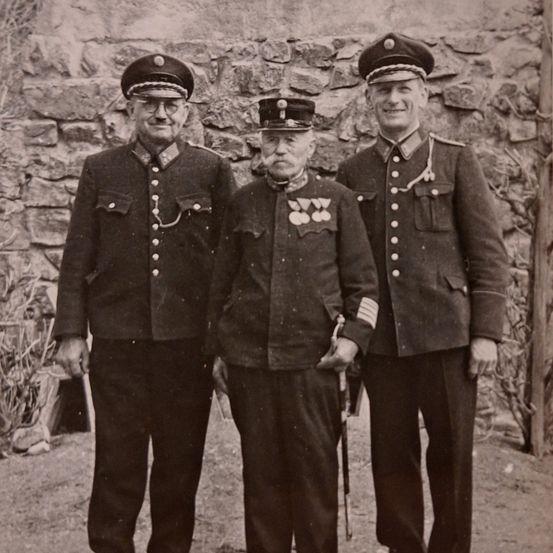 Drei Männer in Uniform stehen vor einer Steinmauer. Sie tragen alle Mützen und Medaillen. Der Mann in der Mitte hält einen Stock.